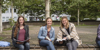 Drei Studentinnen sitzen auf einer Bank, halten Bücher in den Händen und lächeln.